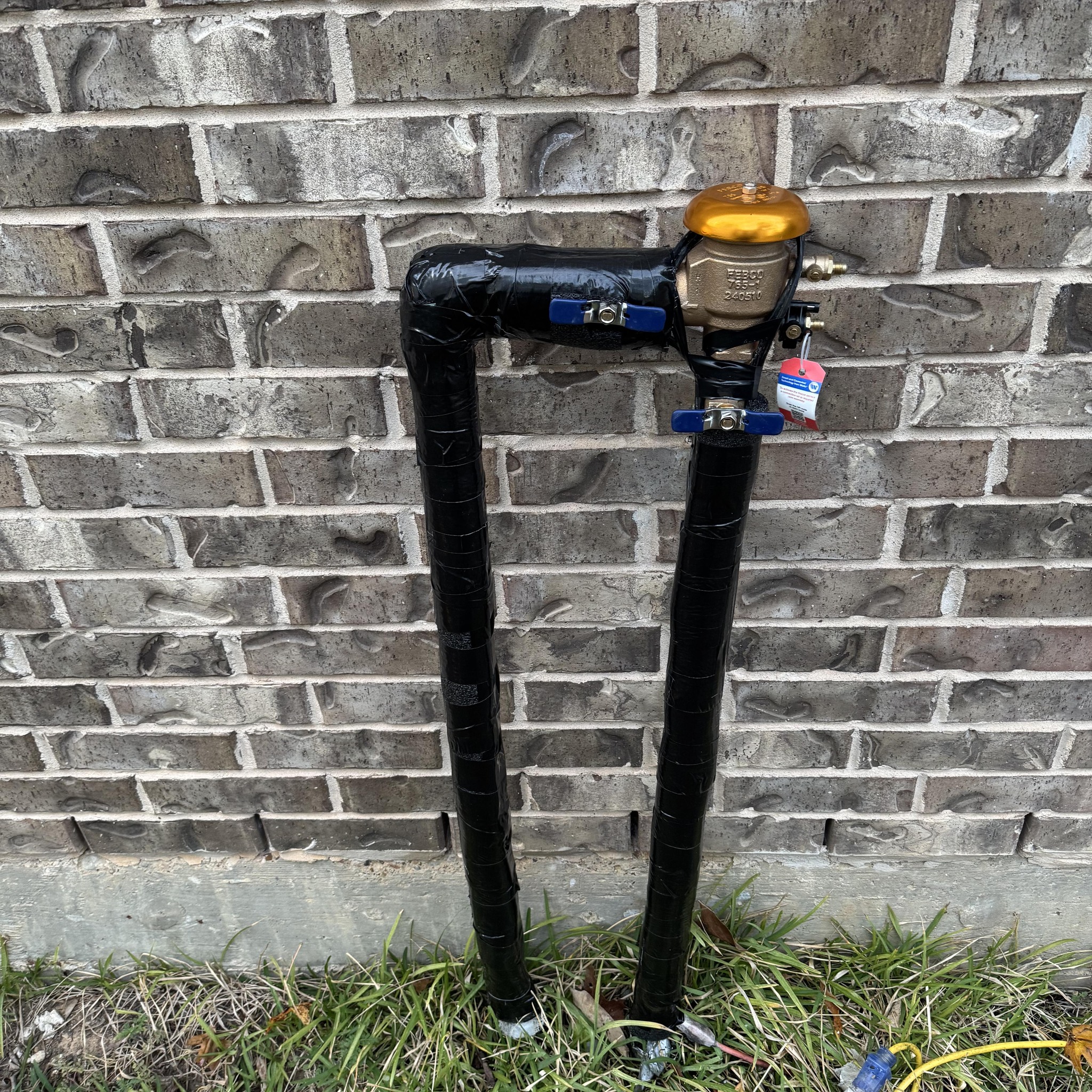 Backflow prevention assembly with insulated pipes installed on the exterior wall of a brick home, used for annual backflow testing and protection of the clean water supply in Conroe, Texas.