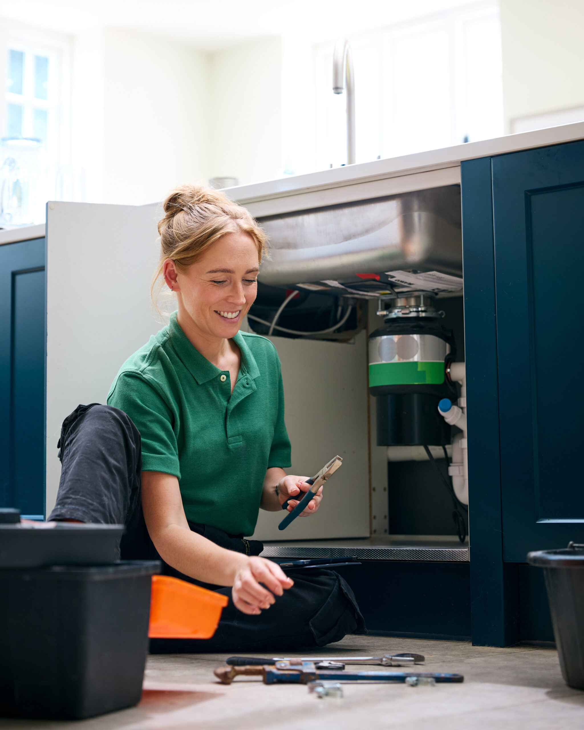 Female plumber installing or repairing a garbage disposal unit under a kitchen sink, using tools and wearing a green uniform during a residential plumbing service in Conroe, Texas.